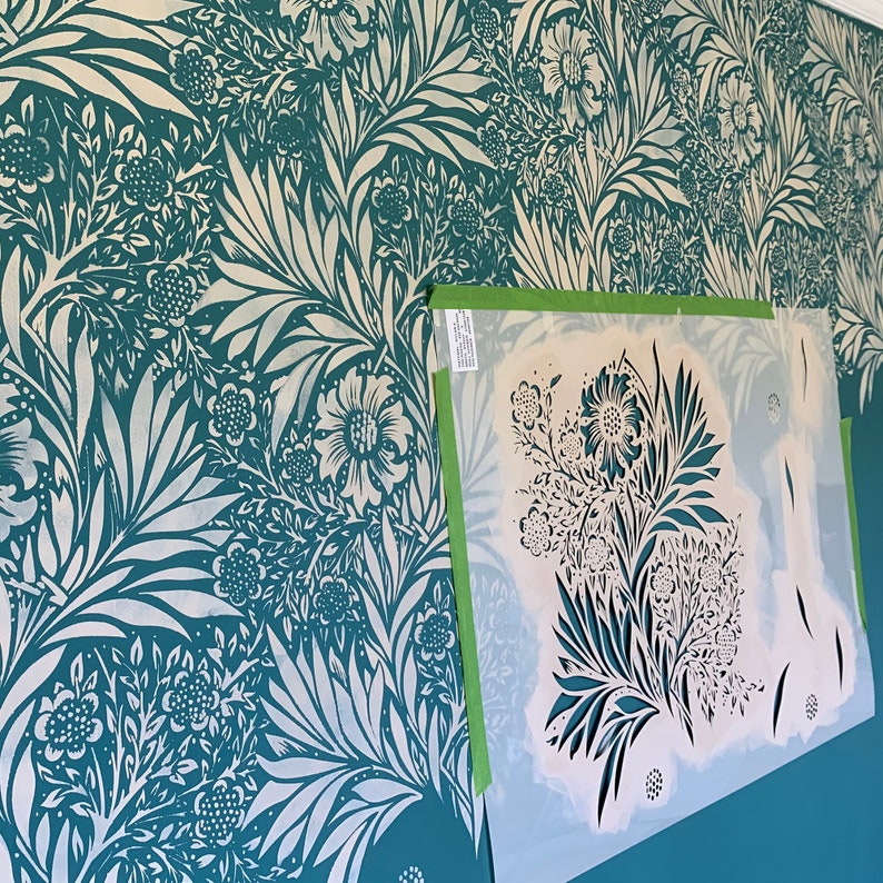 May include: A teal green wall with a white stencil design of a floral pattern. The stencil is partially applied to the wall, and the design is visible through the stencil.