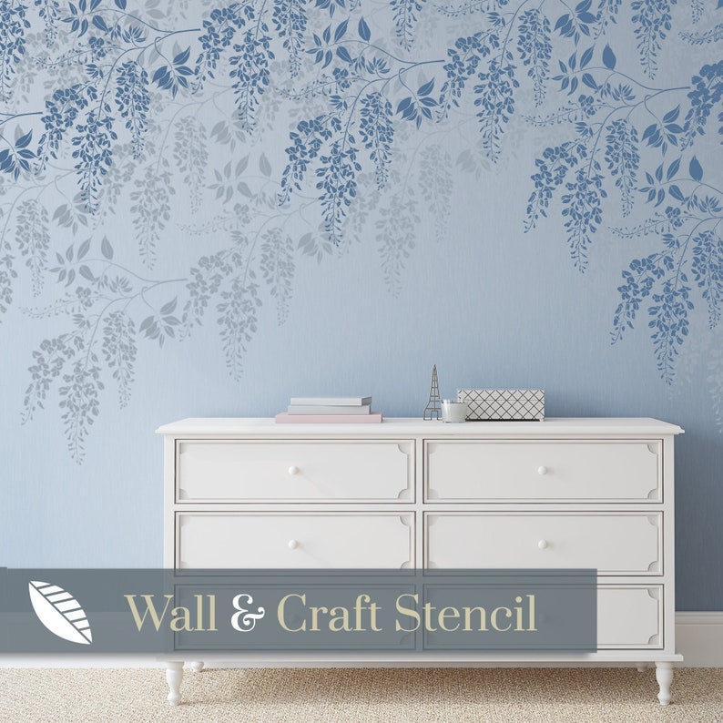 Pu&ograve; includere: Uno stencil da parete con un delicato design di fiori e foglie di glicine. Lo stencil &egrave; realizzato in plastica resistente ed &egrave; riutilizzabile. Lo stencil &egrave; mostrato su una parete blu chiaro con una cassettiera bianca in primo piano. Il testo "Wall & Craft Stencil" &egrave; visibile nella parte inferiore dell'immagine.