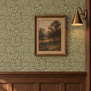 May include: A sage green wallpaper with a repeating cream-colored vine and leaf pattern. A framed landscape painting hangs on the wall, depicting trees and a path. A brass wall sconce with a conical shade is mounted to the right of the frame.