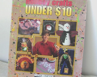 May include: A softcover book titled holiday crafts UNDER $10 from the CLEVER CRAFTER SERIES. The tan cover features colorful paint splatters and multiple inset photographs showing various seasonal handmade projects including a snowman and witch.