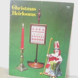 May include: A softcover craft book titled Christmas Heirlooms Book II with a green cover featuring a red candle in a brass holder, a framed cross-stitch sampler on a wooden stand, and a small figurine of a figure in red and white robes.