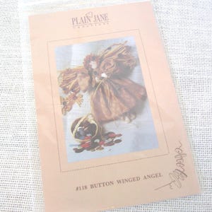 May include: A craft kit featuring a button-winged angel design. The image shows a detailed illustration of an angel with brown wings and a basket of buttons. The text "PLAIN JANE CREATIONS" and "#118 BUTTON WINGED ANGEL" are visible.