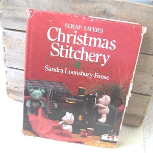 May include: A vintage book titled "Scrap Saver's Christmas Stitchery" by Sandra Lounsbury Foose. The cover is red with white text and features illustrations of teddy bears, a sewing machine, and spools of thread. The book is slightly worn.