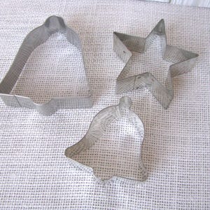 May include: Three metal cookie cutters in the shapes of a bell, a star, and a smaller bell. The cutters are made of a shiny, silver-colored metal and are arranged on a white, textured surface. The bell and star shapes suggest a holiday theme.