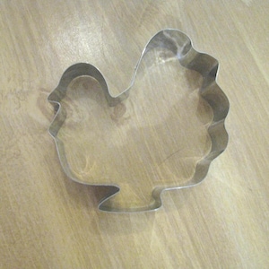 May include: A silver-colored metal cookie cutter shaped like a turkey. The outline features a rounded head, a curved neck, a full body, and a ruffled tail section. The metal edges are smooth and reflective, forming a continuous shape for baking.