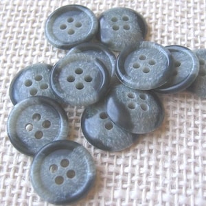 May include: A collection of gray buttons with four holes each. The buttons have a marbled appearance with darker gray edges. They are scattered on a light beige woven fabric.