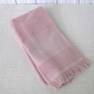 May include: A folded, dusty rose-colored towel with a textured weave and fringed edge. The towel has a solid center panel and a softer, more plush texture on the top and bottom sections. The overall design is simple and classic.