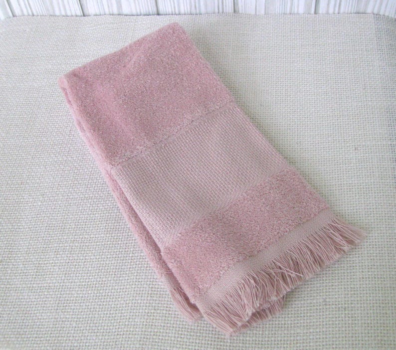 May include: A folded, dusty rose-colored towel with a textured weave and fringed edge. The towel has a solid center panel and a softer, more plush texture on the top and bottom sections. The overall design is simple and classic.