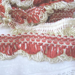 May include: Close-up of decorative trim with a cream-colored base and red accents. The trim features a woven pattern with a scalloped edge of red fringe. This trim is suitable for various craft and sewing projects.