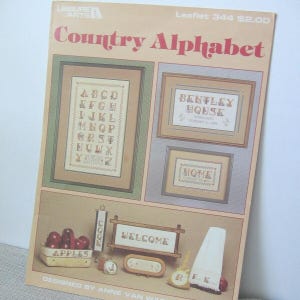 May include: A vintage "Country Alphabet" cross-stitch pattern booklet. The cover features framed cross-stitch samplers with the alphabet, "Bentley House", and "Home". Other images include "Apples" and "Welcome" signs, and a towel with the letters "R.E.K."