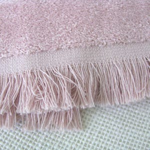 May include: Close-up of a soft, pink towel with a textured surface and a fringed edge. The towel's color is a muted rose, and the fringe adds a decorative element. The towel is made of a plush material.