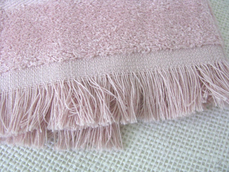 May include: Close-up of a soft, pink towel with a textured surface and a fringed edge. The towel's color is a muted rose, and the fringe adds a decorative element. The towel is made of a plush material.