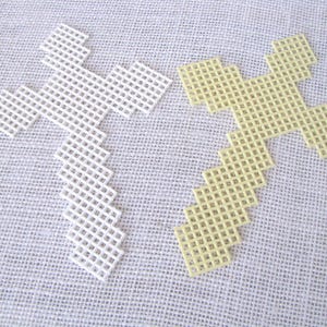 May include: Two plastic canvas crosses, one white and one pale yellow, are displayed on a textured, off-white surface. The crosses are constructed with a grid-like pattern, ideal for needlepoint or cross-stitch projects. The crosses are a craft supply.