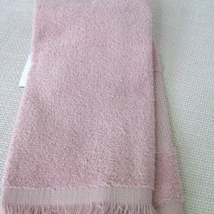 May include: A folded, light pink towel with a textured surface and a fringe detail along the bottom edge. The towel is hanging from a white plastic hanger.