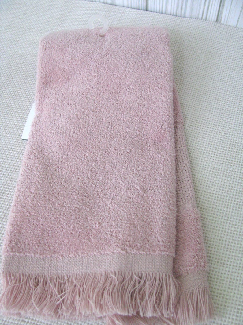 May include: A folded, light pink towel with a textured surface and a fringe detail along the bottom edge. The towel is hanging from a white plastic hanger.