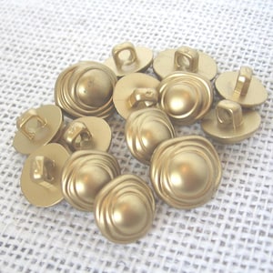 May include: A collection of round, matte gold-toned buttons featuring a raised, concentric circular design on the front and a sturdy shank loop on the back for attachment. These metallic-finish buttons have a smooth, uniform surface texture.