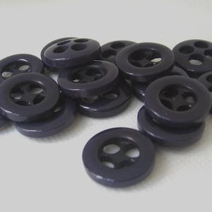 May include: A collection of round, dark purple buttons, each featuring a smooth, glossy finish and four central holes arranged in a square pattern. These circular fasteners are scattered in a loose pile, showing their uniform shape and deep color.