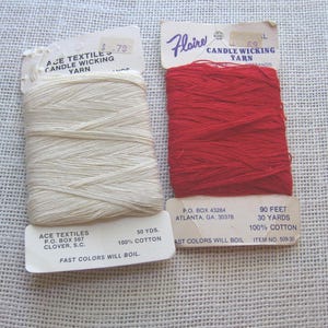 May include: Two spools of candle wicking yarn. One is off-white, labeled "Ace Textiles," and the other is red, labeled "Flaire." Both are 100% cotton. The red spool contains 90 feet of yarn, while the off-white spool contains 50 yards.