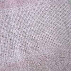 May include: Close-up of a light pink towel with a textured weave. The towel features a woven band in the center, bordered by a stitched seam. The bottom and top edges have a soft, plush texture.