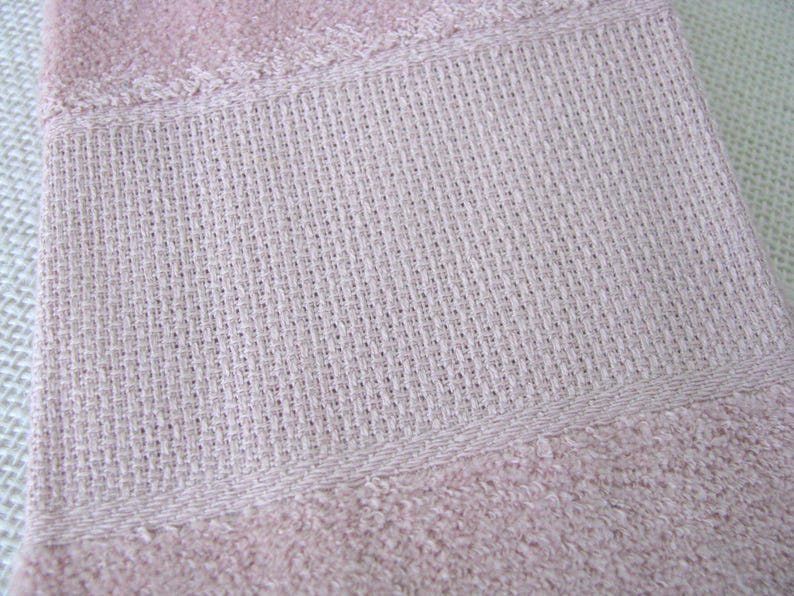 May include: Close-up of a light pink towel with a textured weave. The towel features a woven band in the center, bordered by a stitched seam. The bottom and top edges have a soft, plush texture.