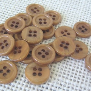 May include: A collection of light brown, round buttons with four holes each. The buttons are made of a smooth, possibly plastic material, and are scattered on a textured, off-white surface. The buttons vary slightly in color and size.