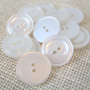 May include: A collection of white, pearlescent buttons. The buttons have two or four holes and a slightly raised rim. The buttons are round and vary in size. The buttons are scattered on a textured, beige surface.