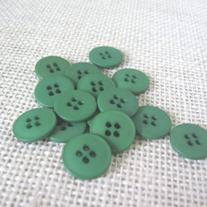 May include: A collection of round, green buttons with four holes each. The buttons are scattered on a textured, off-white surface. The buttons are a solid, matte green color.
