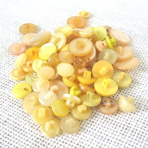 May include: A pile of assorted yellow buttons in various shapes and sizes. The buttons range in color from pale yellow to a deeper, golden hue. Some have two or four holes, while others have a shank. The buttons are made of plastic.