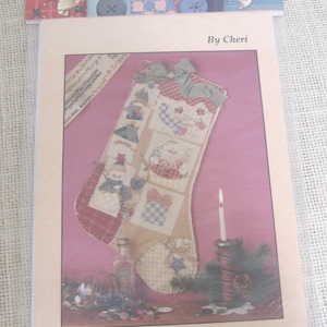 May include: A craft pattern packet titled Hung by Chimney by Cheri, featuring a quilted stocking design with fabric patches, a green gingham bow, and decorative buttons. The cover displays a finished stocking, a candle, and loose buttons.