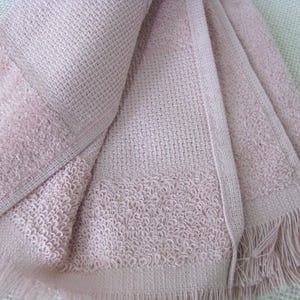 May include: A folded, light pink woven towel with a textured pattern and fringe detail. The towel has a waffle-like weave in the center and a more textured border. The fringe is along the bottom edge.