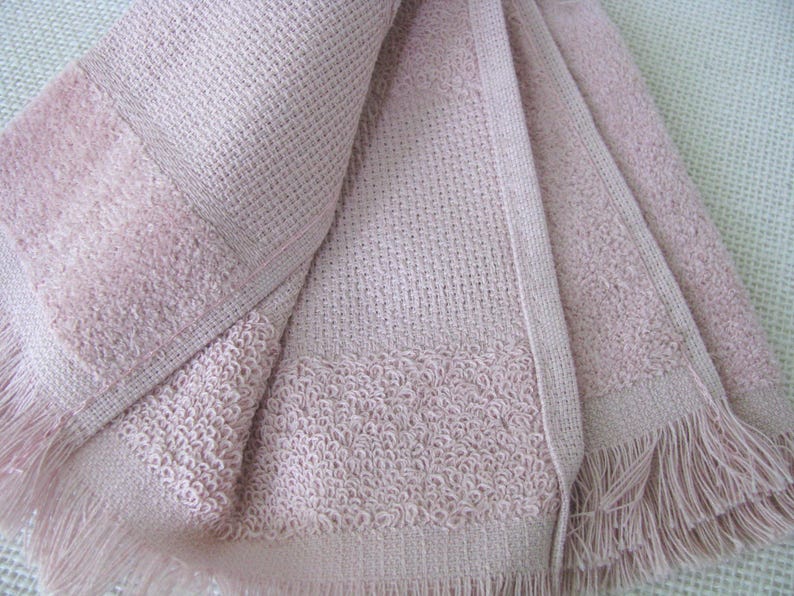 May include: A folded, light pink woven towel with a textured pattern and fringe detail. The towel has a waffle-like weave in the center and a more textured border. The fringe is along the bottom edge.