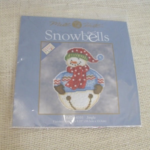 May include: A Mill Hill Snowbells cross stitch kit featuring a snowman wearing a red and green hat, a red polka dot scarf, and a blue sweater. The packaging displays the text Debbie Mumm, Snowbells, and Jingle, with a finished size of 4 x 5.25 inches.