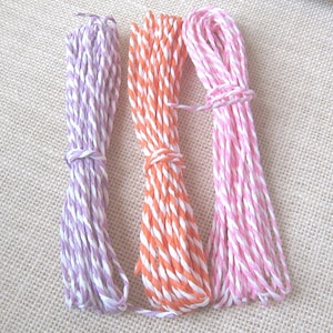 May include: Three bundles of twisted twine in shades of purple, orange, and pink with white accents. Each bundle is tied with a matching twine. The twine is likely used for crafting, gift wrapping, or decorative purposes.