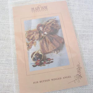 May include: A craft pattern packet titled Plain Jane Creations #118 Button Winged Angel. The cover features a photograph of a brown fabric angel figure with wings and a small basket filled with various colorful buttons on a white surface.