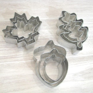 May include: Three sets of silver-colored metal cookie cutters, each containing multiple nested pieces. The shapes include a maple leaf, an oak leaf, and an acorn. These kitchen tools are constructed from thin, polished metal strips formed into outlines.