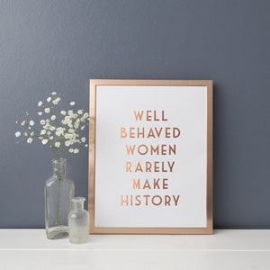 Pode incluir: Uma impress&atilde;o emoldurada com o texto "WELL BEHAVED WOMEN RARELY MAKE HISTORY" em letras cor de cobre. A moldura &eacute; cor de cobre. Um vaso de vidro transparente com flores brancas e uma garrafa de vidro pequena.