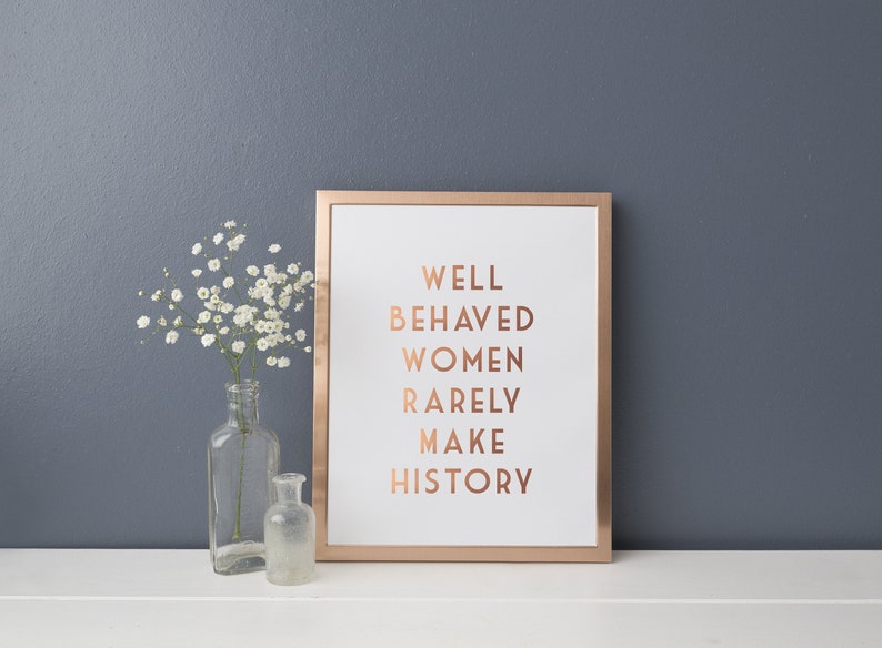 Pode incluir: Uma impress&atilde;o emoldurada com o texto "WELL BEHAVED WOMEN RARELY MAKE HISTORY" em letras cor de cobre. A moldura &eacute; cor de cobre. Um vaso de vidro transparente com flores brancas e uma garrafa de vidro pequena.