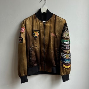 May include: A gold satin bomber jacket with a black zipper and black ribbed cuffs and waistband. The jacket has several patches sewn on the left sleeve, including a Harley Davidson patch, a Daytona 500 patch, and several HOG patches.