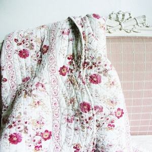May include: A quilted throw blanket featuring a white base with a repeating floral pattern in shades of deep pink, muted red, and soft beige. The fabric displays vertical bands of intricate botanical motifs and quilted stitching lines.