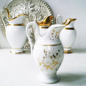 Könnte beinhalten: Vier weiße Porzellankrüge mit goldenen Verzierungen und Akzenten. Die Krüge variieren in Form und Größe, einer davon mit floralen Golddetails. Die Krüge sind auf einer weißen Oberfläche angeordnet, was auf ein Vintage- oder antikes Thema hindeutet.