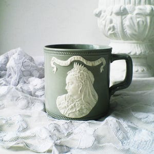 Può includere: Una tazza in ceramica grigia con un rilievo bianco del profilo di una donna, forse la regina Vittoria, e uno stendardo con la scritta "Diamond Jubilee 1897". La tazza ha un manico e un bordo scuri, ed è appoggiata su un panno di pizzo bianco.