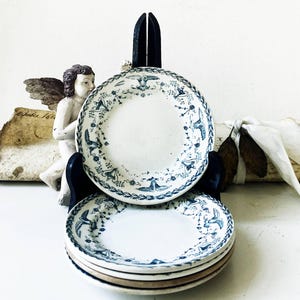 May include: A stack of four round ceramic plates featuring a white base with a blue transferware pattern of floral garlands, urns, and birds. The plates are held upright by a black metal display stand, showcasing the intricate blue decorative border.