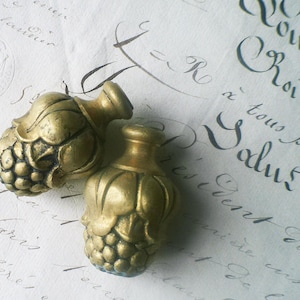 Puede incluir: Dos remates decorativos de color dorado con un diseño de uvas y hojas. Los remates son de metal y tienen un acabado desgastado.