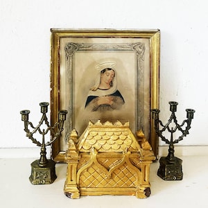 May include: A gold-toned rectangular reliquary box with a scalloped roof, flanked by two dark metal three-arm candelabras. Behind them stands a framed print of a woman with a halo, featuring the text LA VIERGE AU VOILE at the bottom.