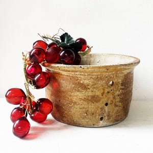 May include: A cylindrical tan ceramic vessel with a textured, mottled surface and three small circular holes on the side, paired with a decorative cluster of translucent, deep red glass grapes featuring green plastic leaves and a vine stem.