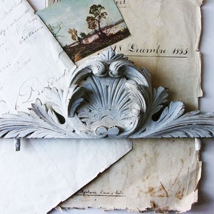 Puede incluir: Un elemento arquitectónico de madera tallada, de color blanco roto desgastado, con un diseño simétrico de hojas estilizadas y un motivo central en forma de concha. Descansa sobre papel envejecido con escritura manuscrita y una pequeña pintura de paisaje.