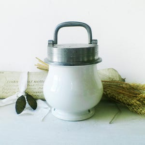 Pode incluir: Jarro de cerâmica branca com tampa de prata e alça. O jarro tem um corpo arredondado e uma base plana. A tampa tem um design decorativo.