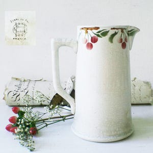 Op de afbeelding: Crèmekleurige keramische kan met een handvat en tuit. De kan is versierd met een kersenboomtakontwerp in rood en groen. De kan heeft een craquelé-glazuur en een stempel met "France".