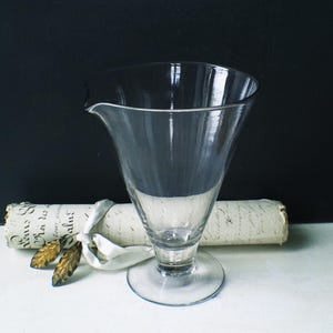 May include: A clear, vintage glass with a flared rim and a small spout. The glass sits on a round base. A rolled parchment with text is behind the glass, tied with a white ribbon and gold leaves.