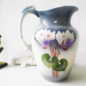 May include: A vintage ceramic jug with a blue and white glaze. The jug features a floral design with pink and purple cyclamen flowers and green leaves.